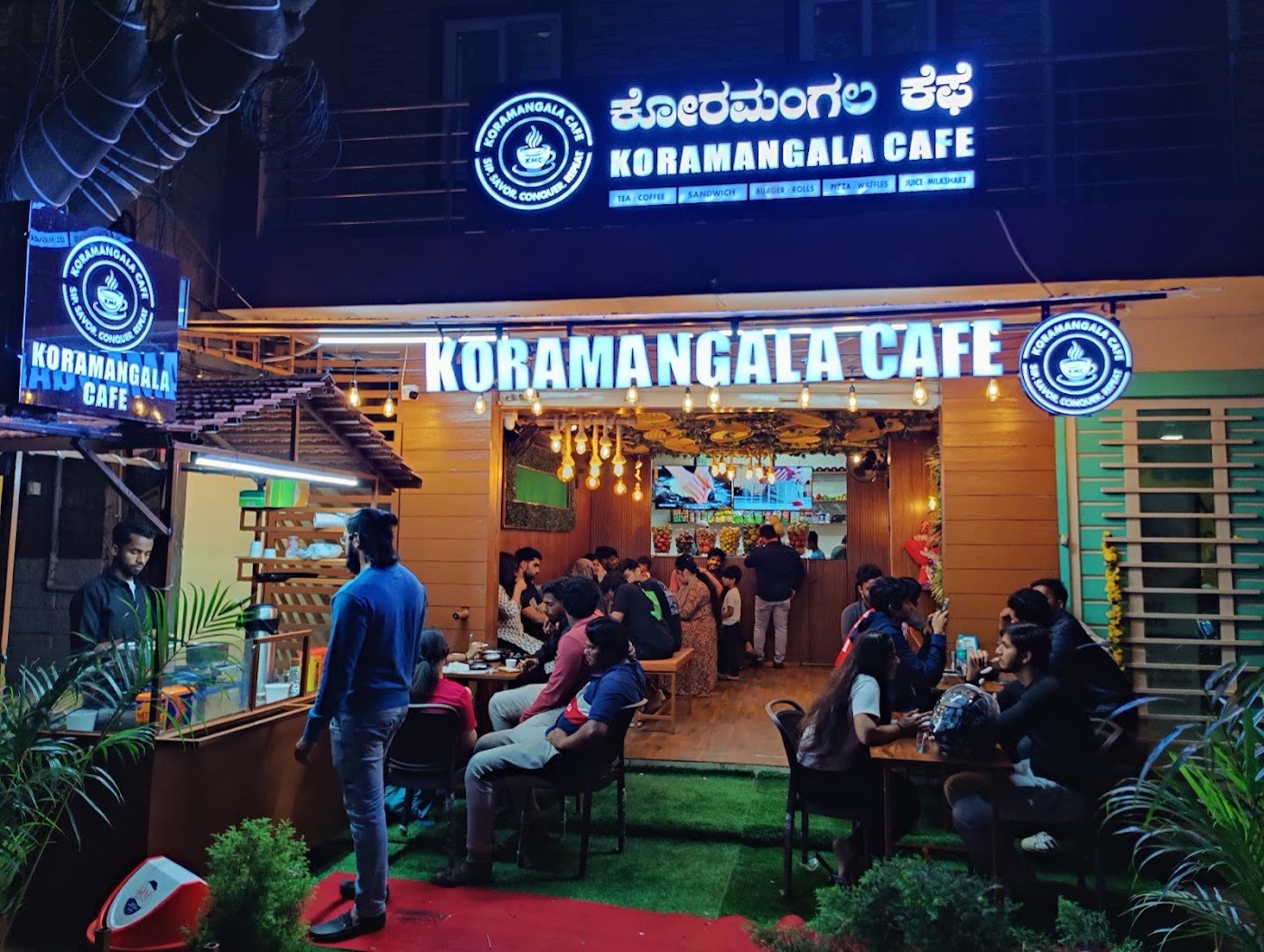 Koramangala Cafe lit up at night with a blue neon sign and people at the tables